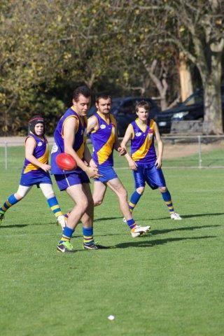 4 participants wearing purple and gold football jerseys, a red Aussie rule football is bouncing towards one player