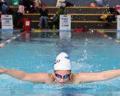 A young female swimmer in a white cap and goggles performs a butterfly stroke in a competitive swimming pool, with spectators in the background.