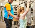 A trainer assists a woman with her workout at a gym, guiding her during a strength training exercise on a machine.