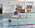 A busy indoor swimming pool, featuring children and adults swimming and playing, with colorful lane markers and a lifeguard on duty.