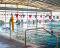 Indoor swimming pool with clear blue water, red lane markers, and swimmers enjoying the space under a spacious, sunlit roof.