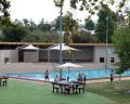 A swimming pool area with children playing, surrounded by greenery, picnic tables, and shaded seating under umbrellas.