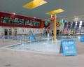 Indoor swimming pool featuring colorful decorations, shallow water areas, and safety signs, under a modern ceiling with large windows.