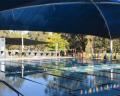 A serene outdoor swimming pool covered by blue shade sails, surrounded by trees and seating areas. Water glistens under sunlight.