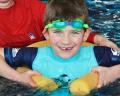 A child in a light blue swim vest is being supported by an adult in a red shirt, both in a swimming pool.