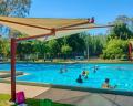 A sunny outdoor pool scene with children swimming and adults supervising under shaded canopies and surrounded by lush greenery.