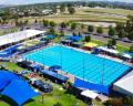 Aerial view of a large swimming pool complex surrounded by blue tents, lush grass, and distant hills, bustling with activity.