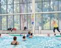 An instructor leads a water aerobics class in a bright indoor pool, surrounded by participants in the water.