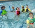 A group of children and an instructor enjoy swimming in a pool, using colorful pool noodles for support and safety.