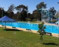 A sunny outdoor swimming pool with lanes, diving boards, shaded areas, and landscaped greenery surrounding it.