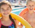 Three children, two girls and a boy, stand together with colorful pool noodles at a sunny swimming pool.