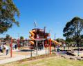 A vibrant public playground featuring a slide, climbing structures, and a splash pad with children playing under a clear blue sky.