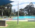 A panoramic view of an outdoor swimming pool surrounded by lush green grass and trees under a partly cloudy sky.