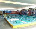 Indoor swimming pool with multiple lanes, bright blue water, yellow railings, and large windows letting in natural light.