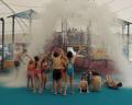 Children in swimwear joyfully gather under a giant water feature, as a torrent of water splashes down in an indoor water park.