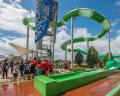 A crowded water park featuring a tall green waterslide, with visitors eagerly waiting in line under a sunny sky.