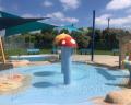 Colorful water play structure with a mushroom cap, spraying water in a sunny park with green benches and shaded areas.