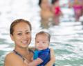 A caregiver holds a baby in a swimming pool, with other adults enjoying the water in the background. The scene is bright and cheerful.