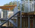 Close-up of a modern play structure featuring a staircase, wooden decks, and wavy metal barriers under a shaded canopy.