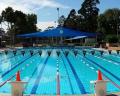 A clear, outdoor swimming pool with lane markers and red cones, shaded by blue canopies, surrounded by green trees under a bright sky.