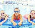 Three children in colorful swimsuits lie on floating mats in a bright, indoor swimming pool, enjoying a fun day in the water.