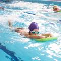 Two children using a green kickboard in the pool, one wearing a purple swim cap.