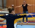 A gymnast balances on a beam while another coach demonstrates proper posture, both focused in a training gym setting.