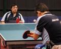 A table tennis game in progress, featuring two players facing each other at a blue table with a ball in motion.