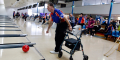 Photo of a tenpin bowling alley, pictured is a man in a blue and red tshirt with black pants and white shoes, holding onto a walker with one hand a bowling a red bowling ball down the lane in the other. You can see other people in the background playing their own games or watching from afar