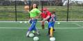 A playful scene on a soccer field features a woman in a bright green shirt, a man in a red cap, and two kids posing with soccer balls.