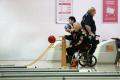 Photo of tenpin bowling lanes with lady in a wheelchair using the accessibility ramp to roll the red bowling ball down the lane. Two other bowlers stand close by.