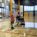 A young boy watches a woman in a wheelchair joyfully tossing a shuttlecock in a brightly lit sports facility.