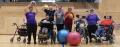 A group of people in purple shirts, some in wheelchairs, joyfully hold exercise balls in a sports hall, promoting inclusivity and teamwork.