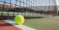 A close-up of a tennis ball resting near the net on a brightly colored court, with a clear blue sky in the background.