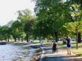 A sunny park scene shows a woman walking a child in a stroller along a tree-lined path beside a calm waterway.