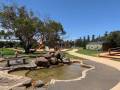 Playground scene featuring a pond, rocky landscape, grassy areas, trees, slides, and sunny blue skies. Families enjoy outdoor activities.