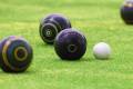 Several colorful lawn bowls in shades of black and blue are positioned on vibrant green grass, with a white jack ball nearby.