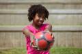 A smiling child with curly hair holds a colorful soccer ball, dressed in a pink shirt, ready to play outdoors.