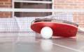 A close-up of a red table tennis paddle resting near a white ping pong ball on a table, with a net in the background.