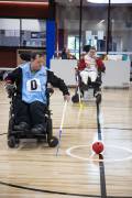 Photo of a man in a wheelchair with a blue jersey on. He is holding a hockey stick and going to hit the ball, another person in a wheelchair can be seen in the background