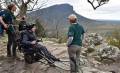 Three people are outdoors on a rocky terrain, one is seated in an all-terrain wheelchair, while two others stand beside, providing assistance. In the background, a mountainous landscape stretches under a cloudy sky.