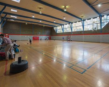 A spacious indoor sports hall with a polished wooden floor, natural light, and players engaged in an activity. Spectators are seated nearby.