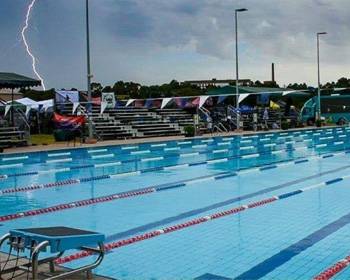 A stormy sky looms over an empty swimming pool, with lightning visible in the background and spectators under tents nearby.